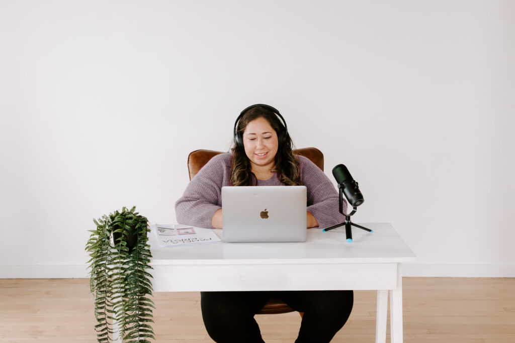 How to Get One Year of Podcast Episode Ideas | Pamela Krista, founder of Moms Who Podcast, wearing a purple shirt at a white desk with a set of headphones recording a podcast episode.